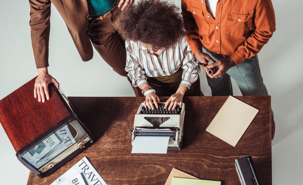 Content writing - lady on typewriter typing and 2 people stood behind watching